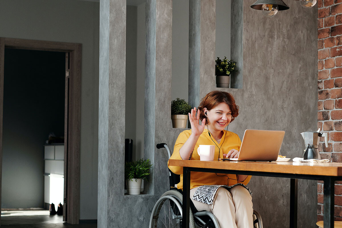 woman in orange top in wheel chair waving to laptop sitting at dining table in modern home