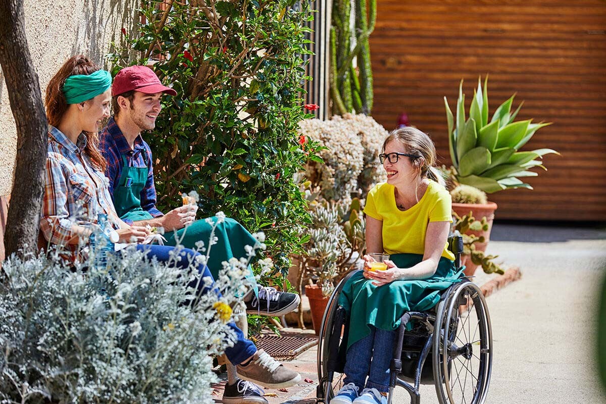Smiling gardeners having food and drink with disabled colleague sitting amidst plants outside greenhouse