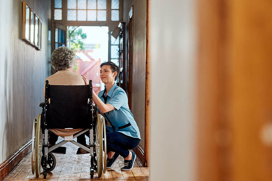 Care working knealing next to older woman in wheelchair in the hallway of a historic home. Open front door in background.