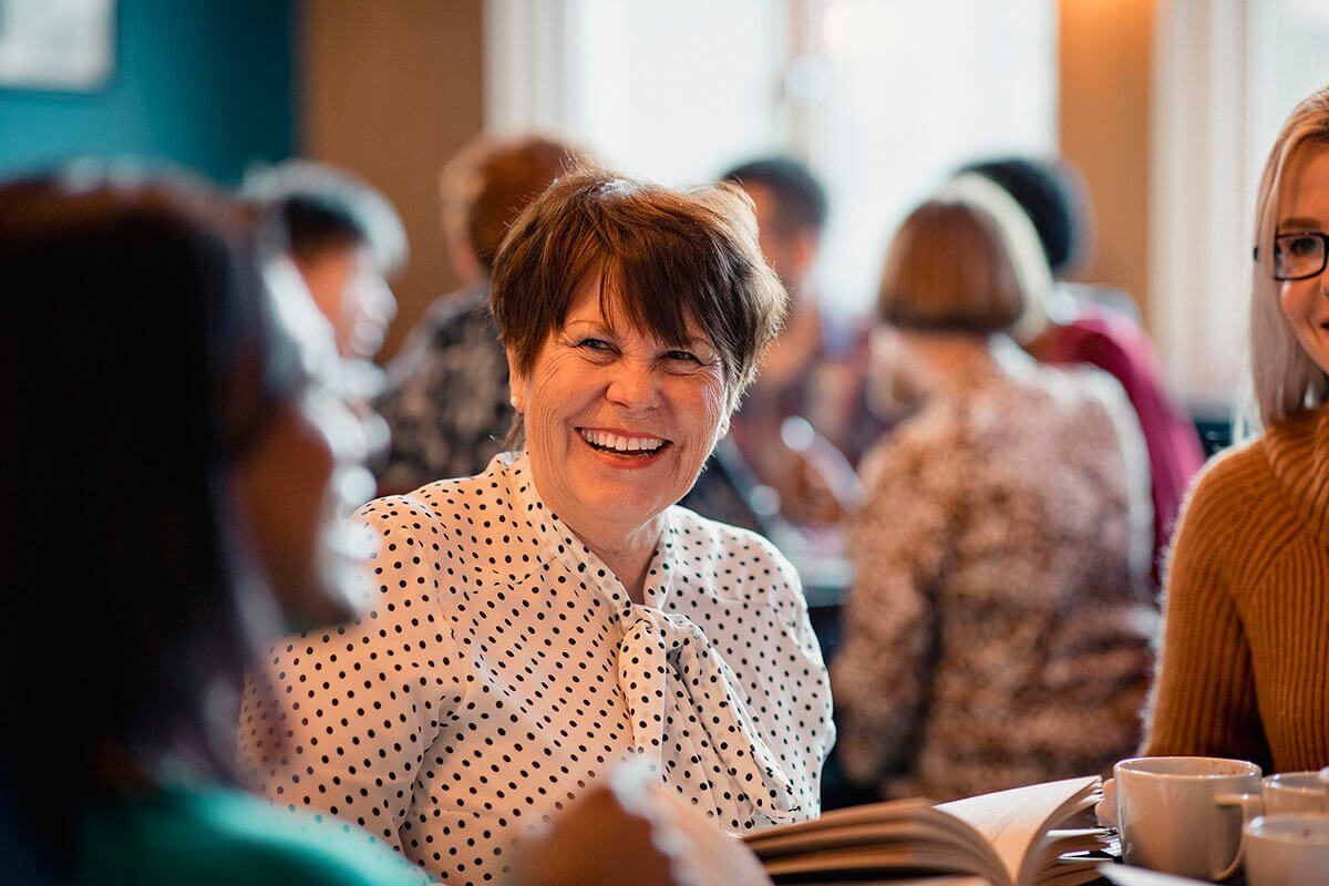 Over the shoulder view of a senior woman laughing and smiling while sitting inside of a cafe. She is enjoying the company of the other women at a conference or meeting